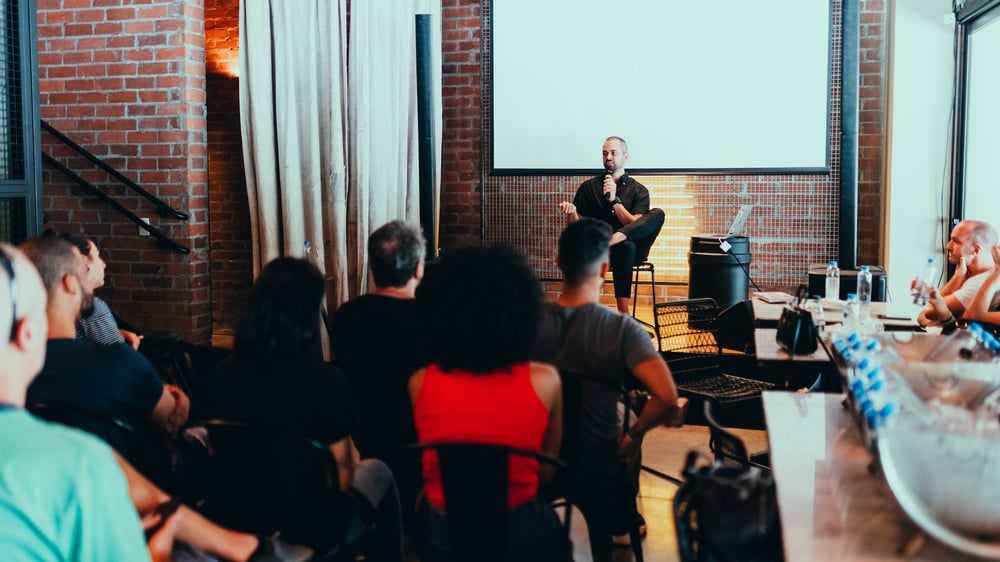 A speaker sits on a stool and talks into a microphone in front of a small crowd in a brick conference room.