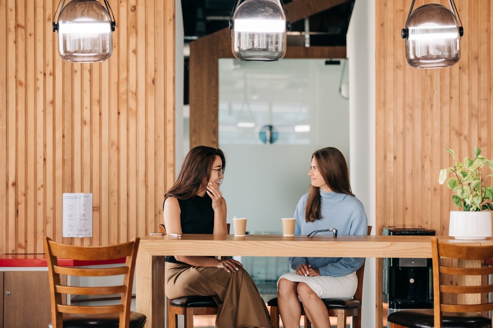 Two people sit at a table in a coffee shop, chatting.