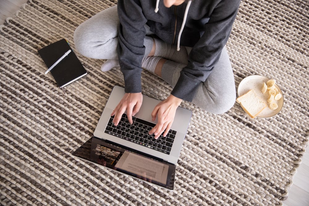 A person wearing sweats sits on the floor and types on a laptop.