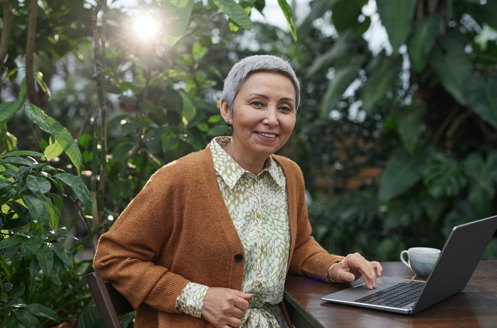 A smiling writer sits in front a laptop at a table surrounded by plants.