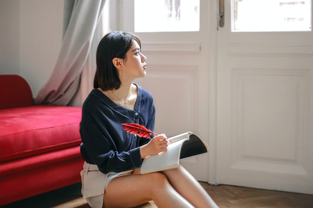 A writer writes in a notebook with a red quill while sitting on the floor and staring out the window.