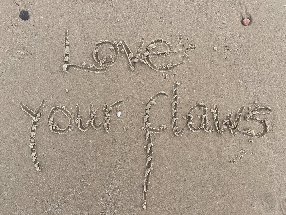 The phrase "Love your flaws" written in the sand.