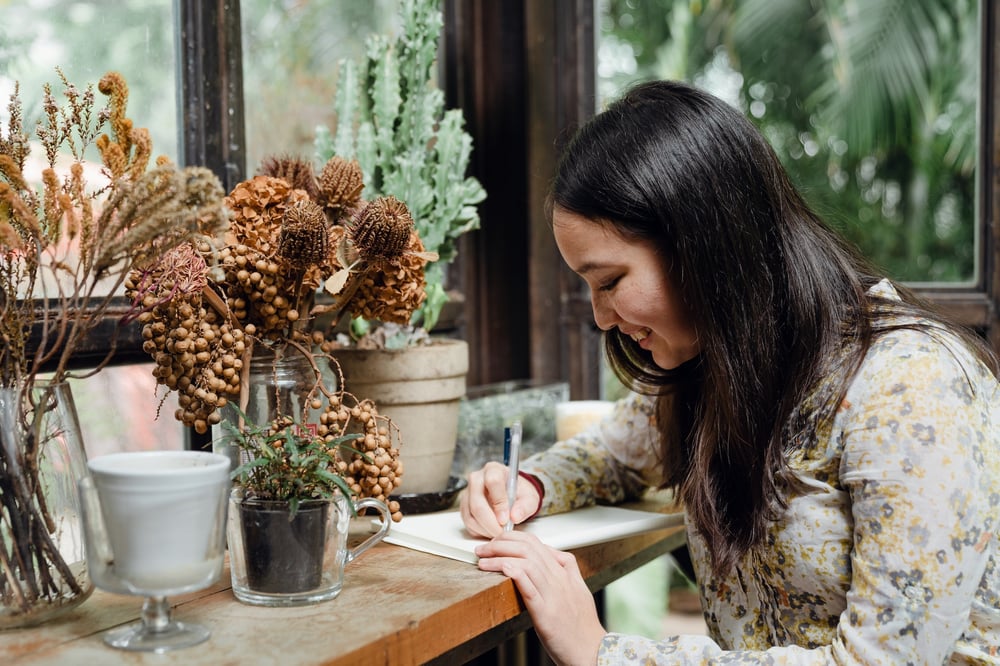 A writer writes in a notebook at a table filled with potted plants.