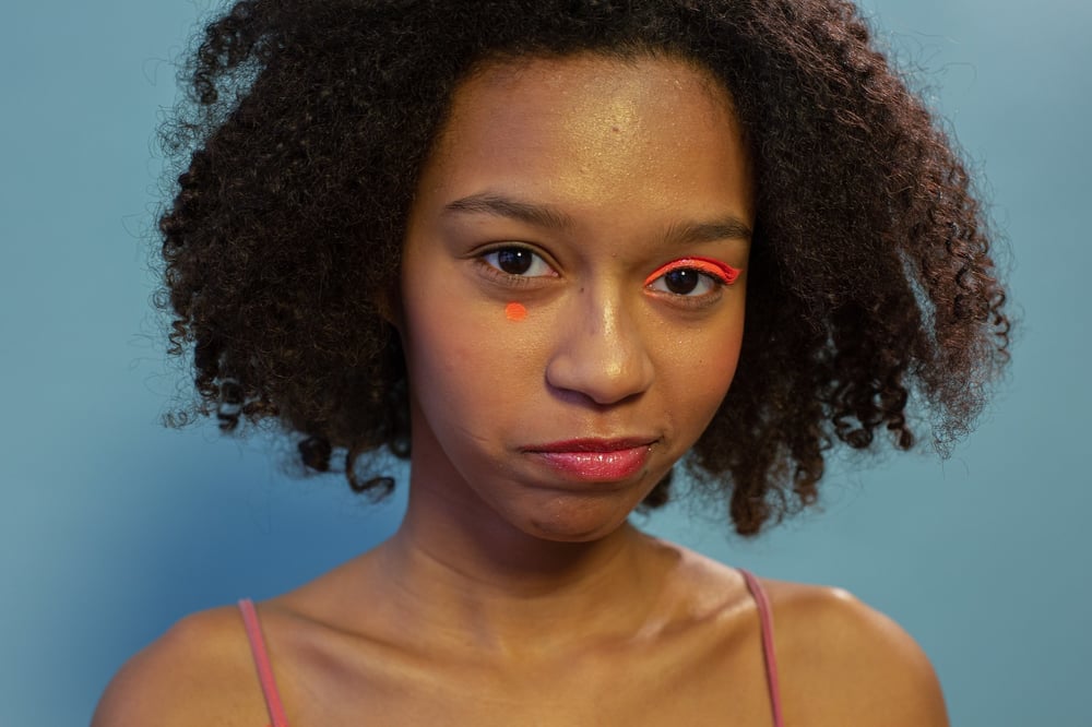 A person with bright eye shadow over one eye smirks.