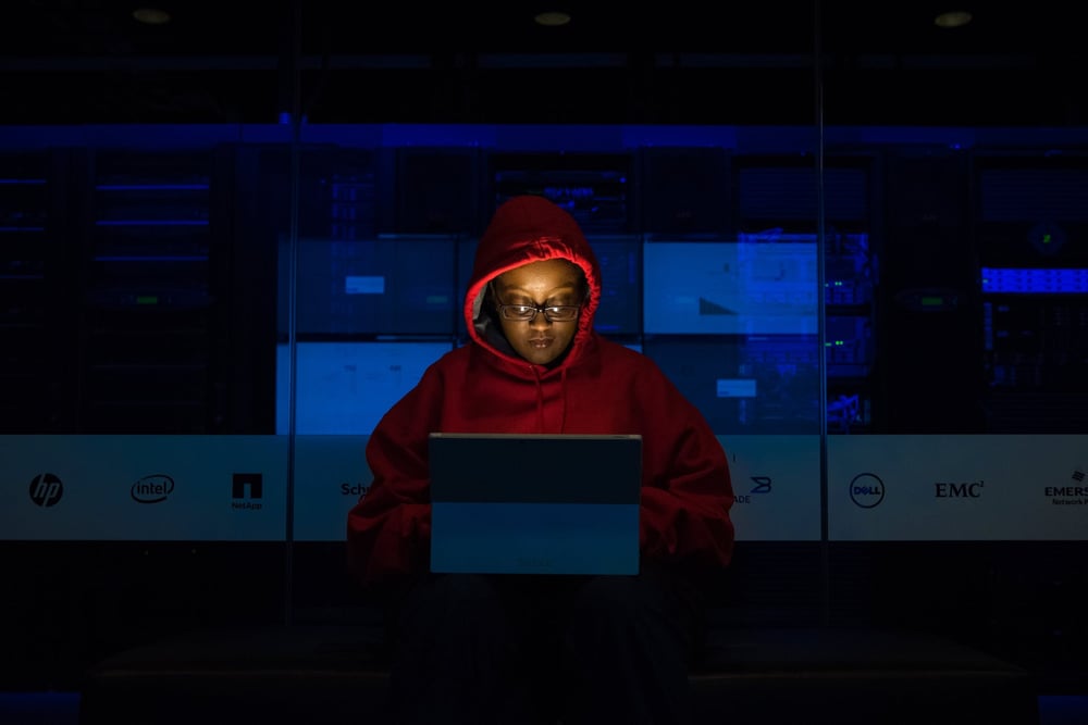 A writer in a red hoodie types on a computer alone in a dark room.