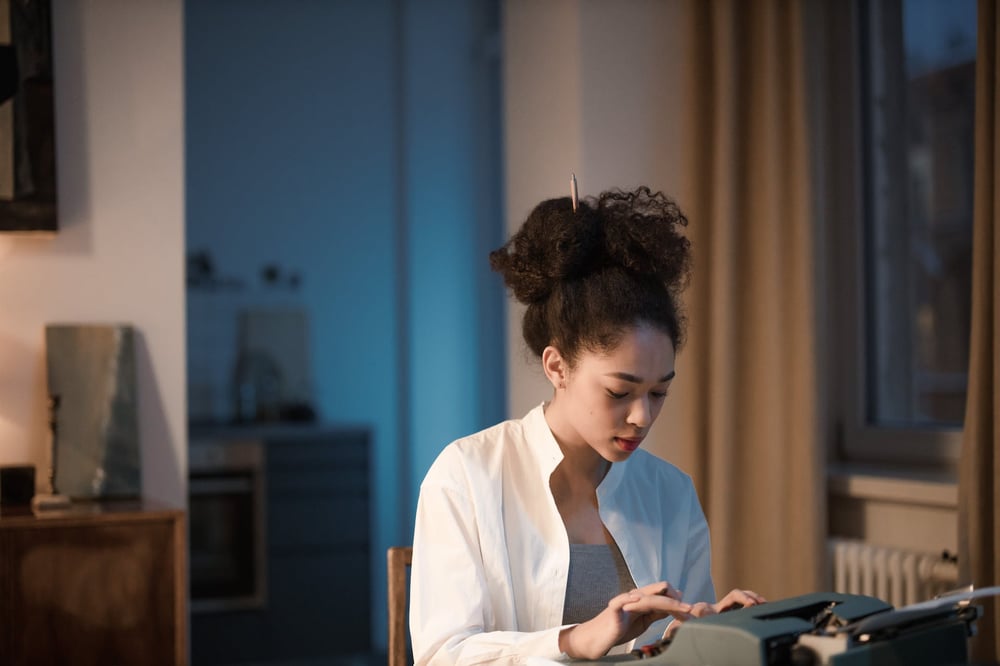 A writer with a pencil in their hair types on a typewriter in a dim room.