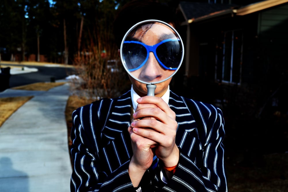 A person in a pin-striped suit and purple sunglasses looks through a magnifying glass.
