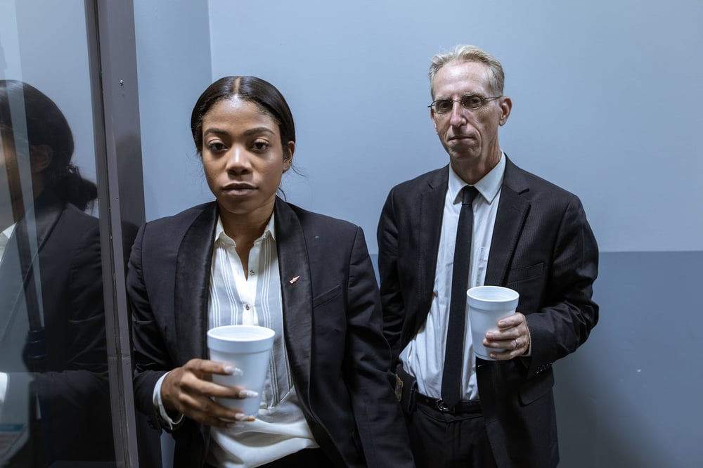 Two detectives stand outside an interrogation room holding coffees.