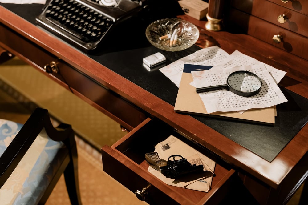 A desk with a gun in an open drawer, a magnifying glass sitting on top of papers with code written on them, a full ashtray, and a typewriter.