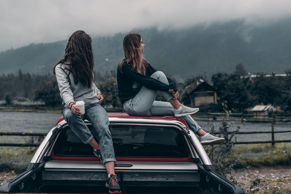 Two teenagers sit together on the roof of a car, looking at a gray sky.