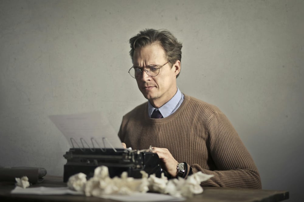 A writer types on a typewriter, surrounded by crumpled pieces of paper.