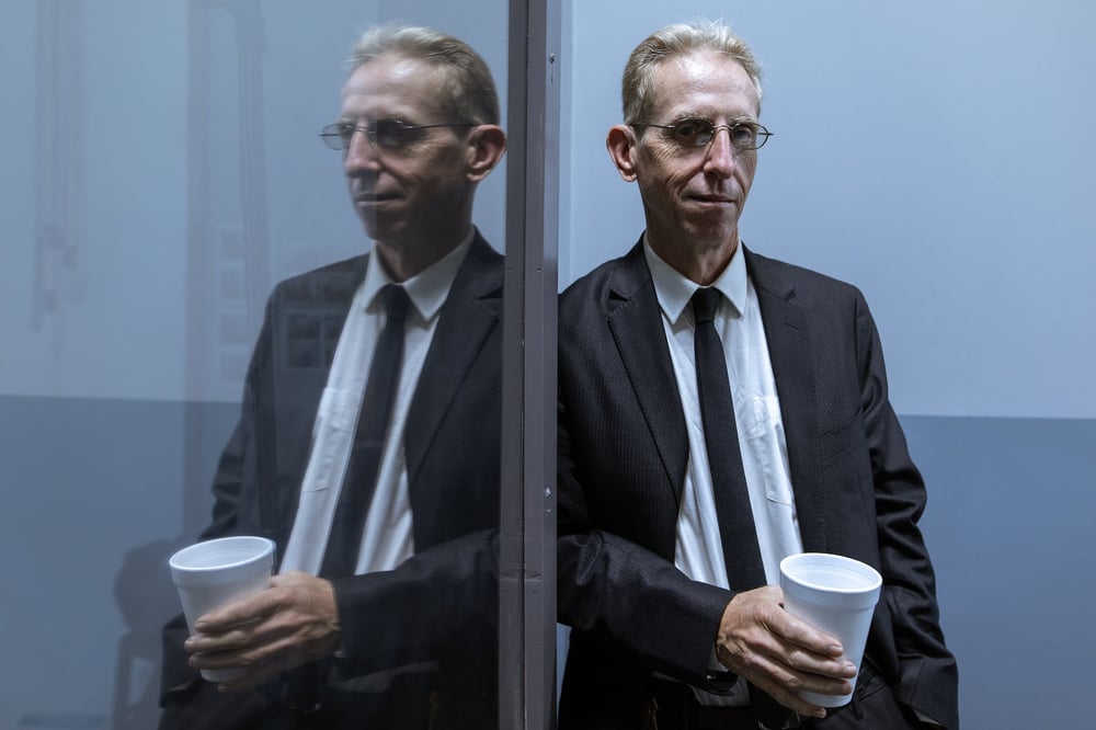 A detective sidekick in a suit and tie holds a cup of coffee and leans against a two-way mirror.