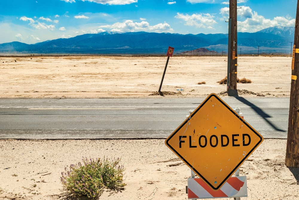 A sign that reads "flooded" sits in a dry desert.