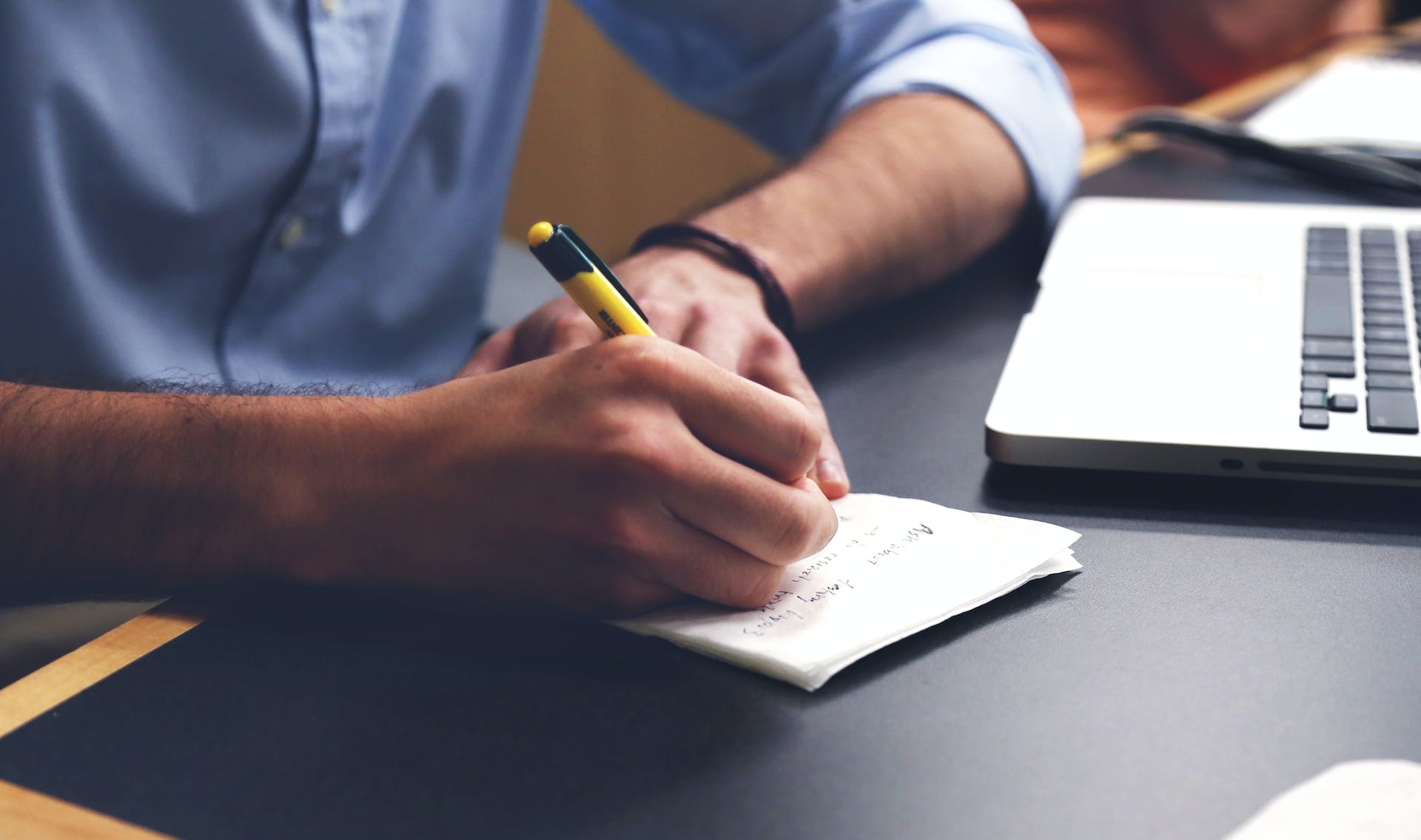 A hand writes on a sheet of paper beside an open laptop.