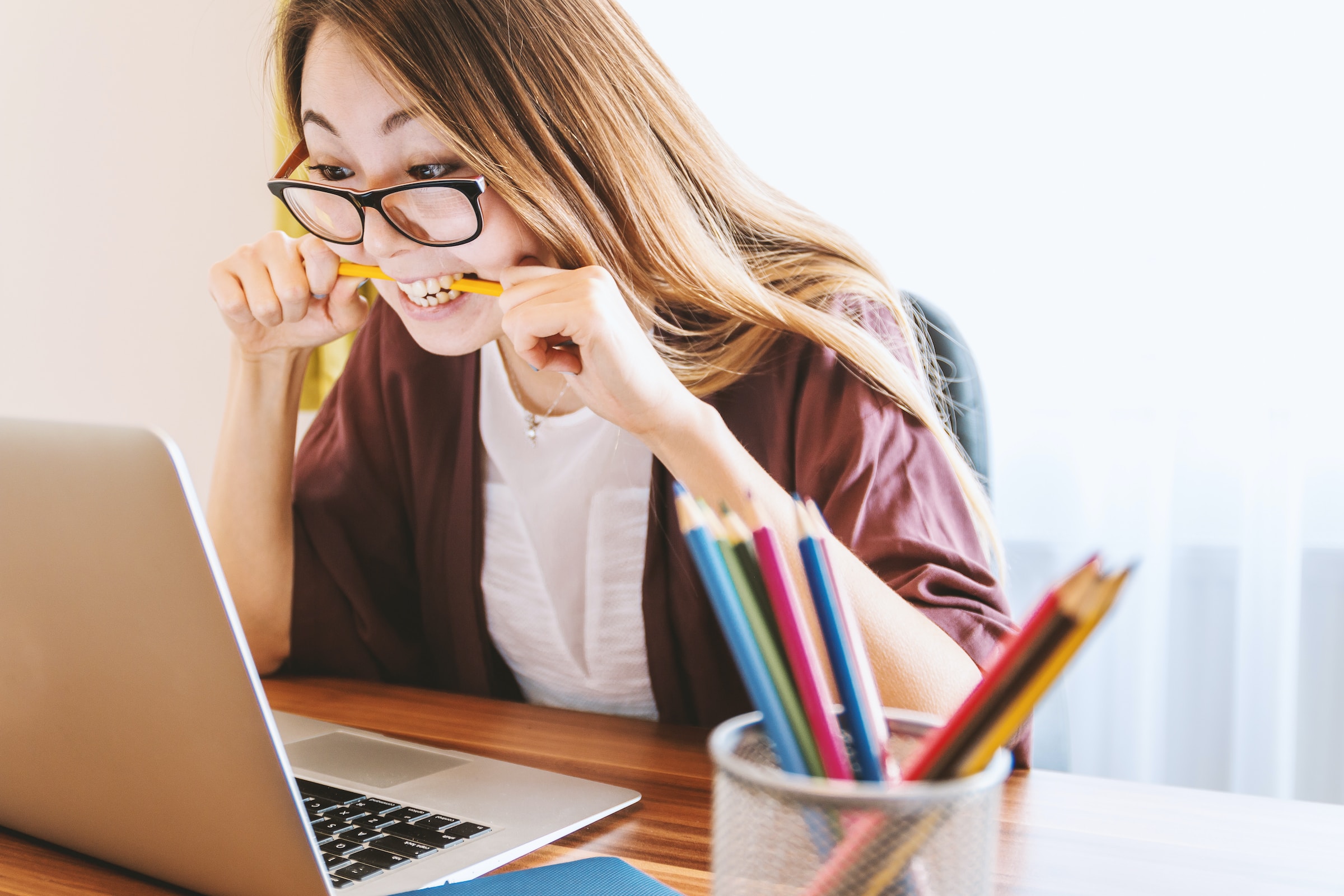 A person wearing glasses looks at a computer screen and bites down on a pencil nervously.