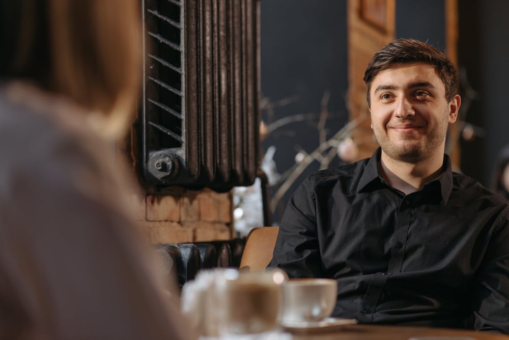 A person sits at a cafe table, smiling at the person across from them.