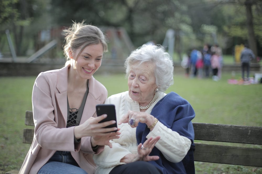 A grandmother and granddaughter sit on a park bench looking at a phone together.