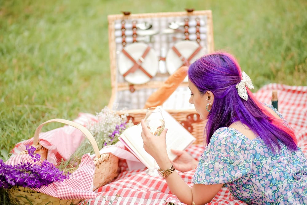 A person with purple hair reads a book on a picnic blanket. A basket of purple flowers sits beside them.