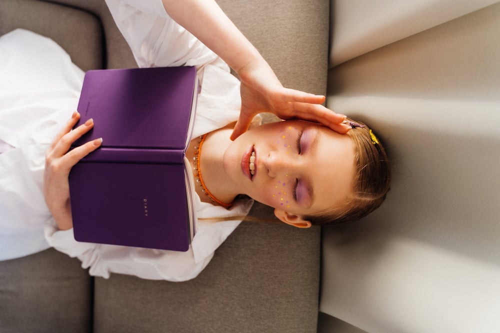 A stressed-looking person touches their hand to their head while lying on a couch, reading a purple book.