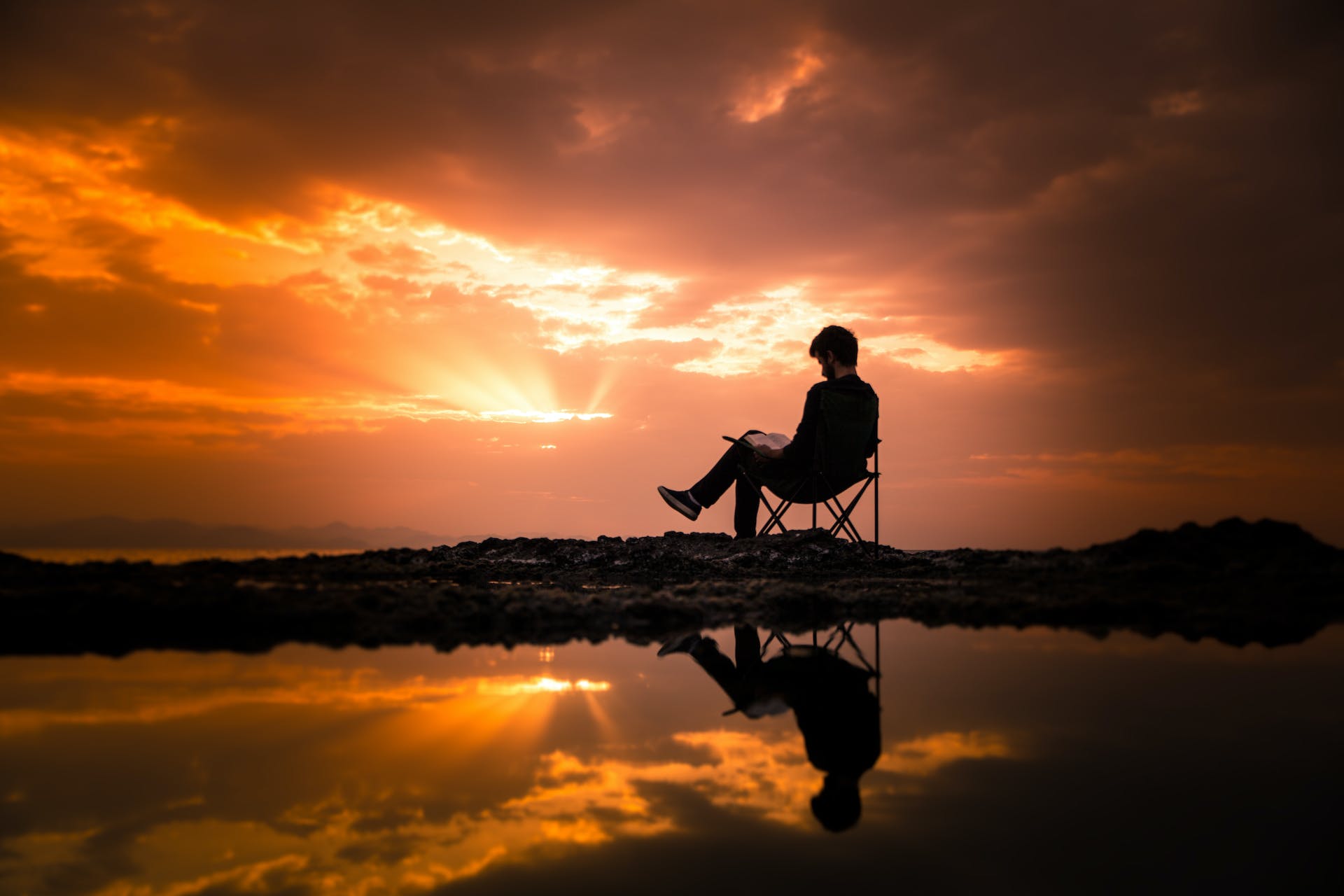 A person sits reading a book in a camping chair on a beach at sunset.