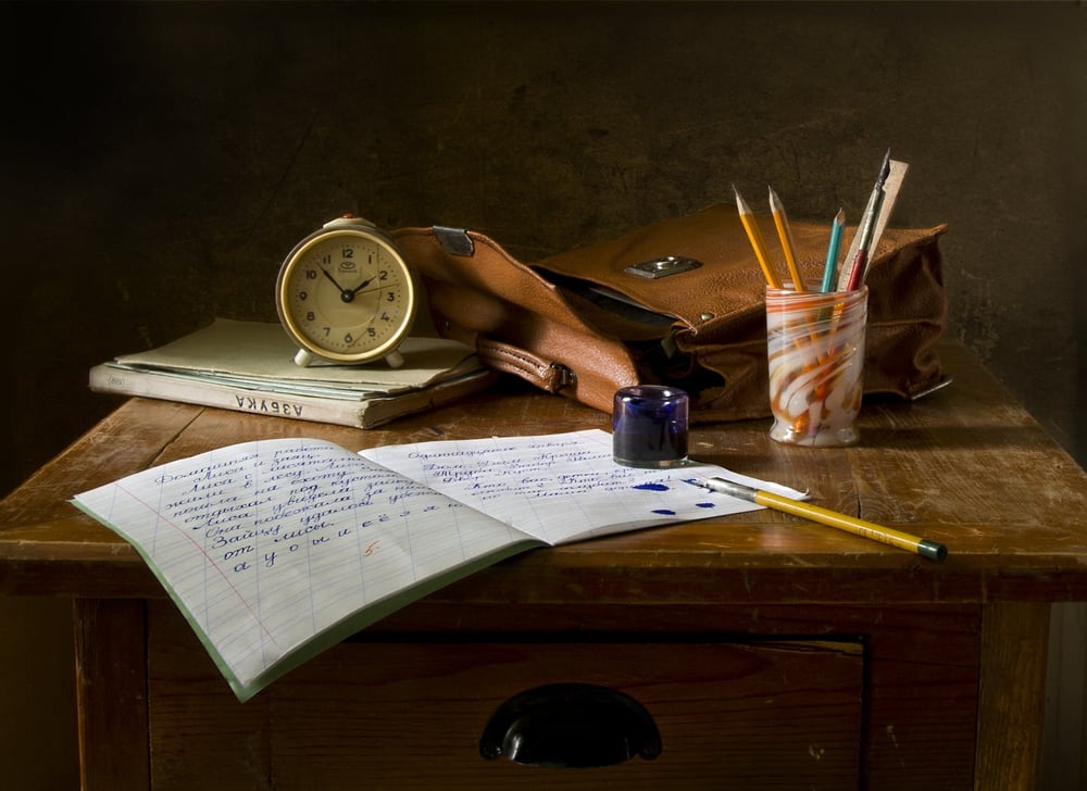 A thin notebook filled with writing and ink smudges its open on a desk beside an ink well, pen cup, clock, books, and leather bag.