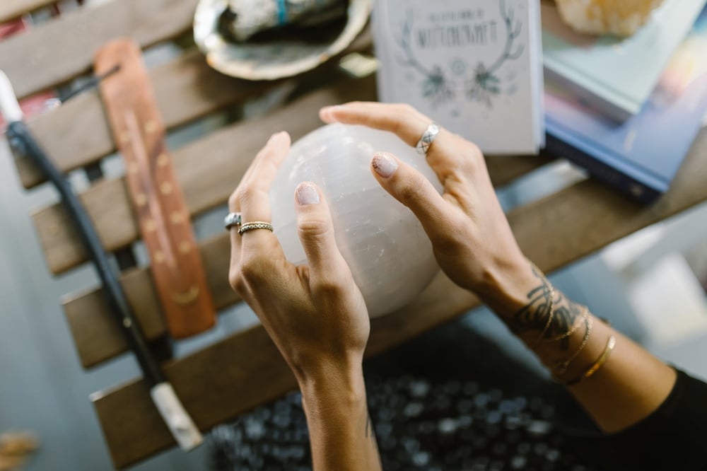 A pair of hands decked out in jewelry palm a crystal ball.