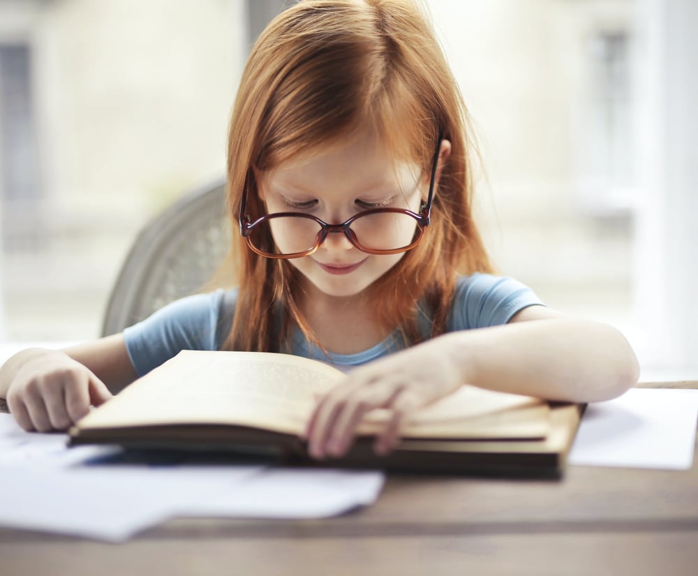 A small child wearing big glasses reads a book at a table.