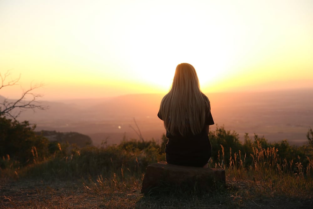 A person sits on a hill at sunset, looking out at the view.