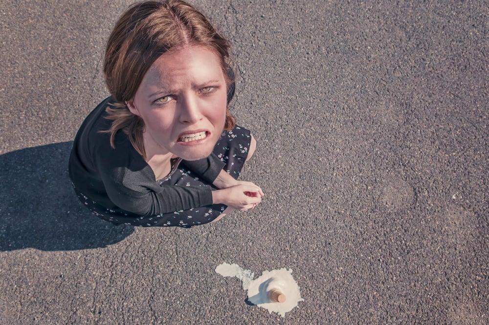 An upset person squats on the ground beside a dropped ice cream cone.