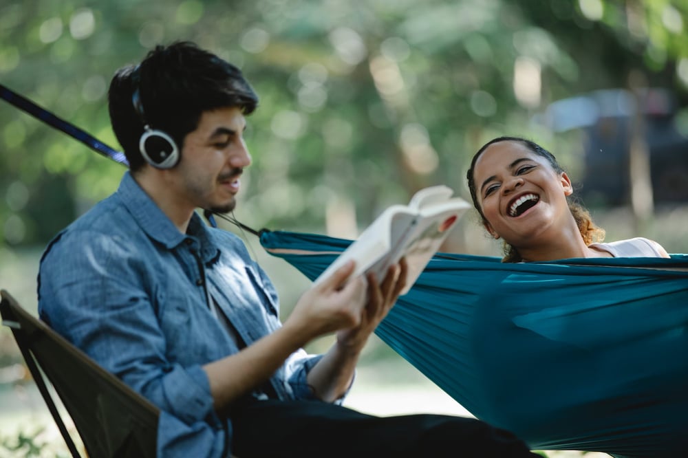 A person lies in a hammock laughing as another person reads to them from a book.