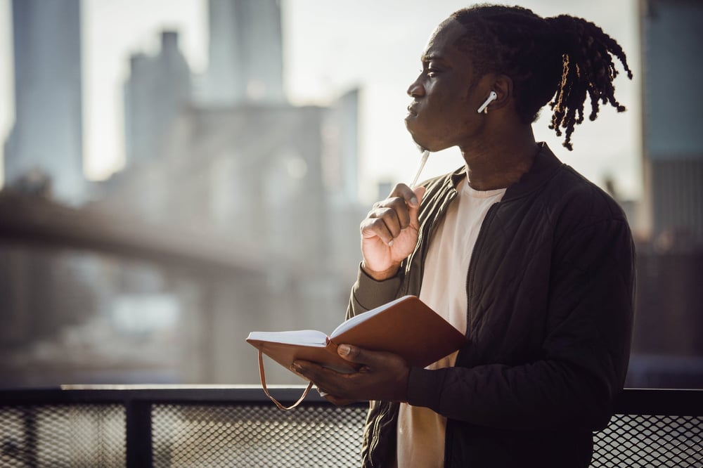 A writer holds an open journal and looks out at a city skyline, touching a pen to their chin and thinking.