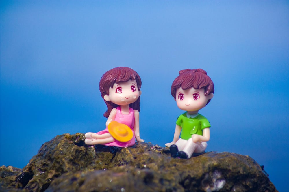 Two toy characters—a boy and a girl with a hat—sit together on a rock.