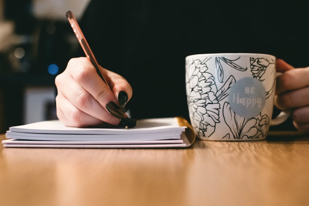 A hand writing in a notebook beside a floral mug that says