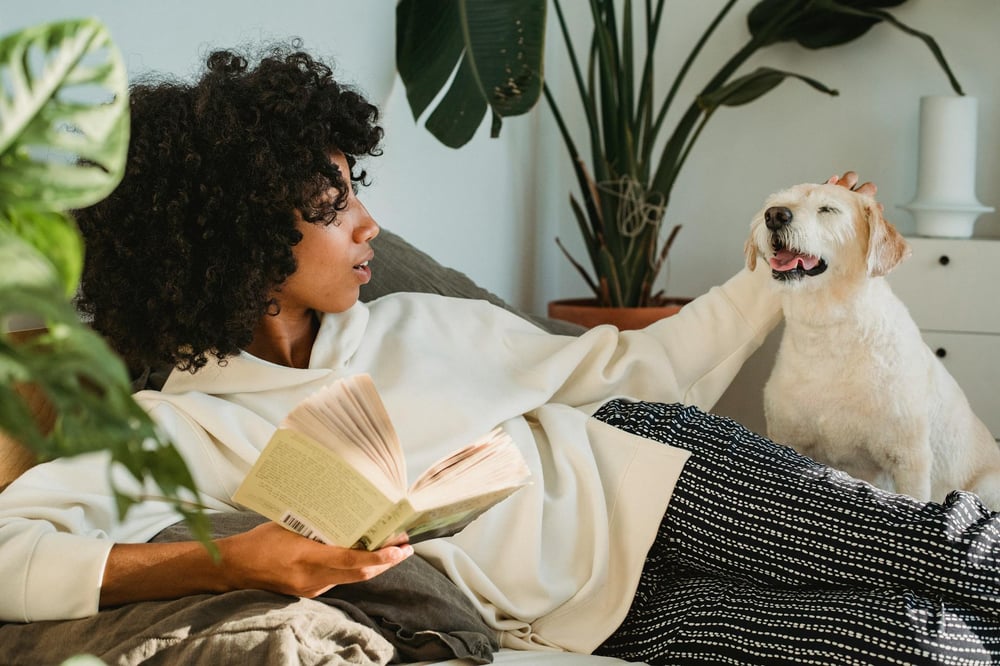 A person lounges on a couch petting a dog and reading a book.