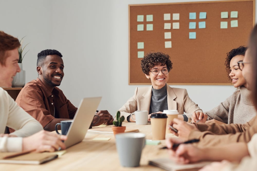 A group of smiling people sit around a table in a meeting room, surrounded by coffee cups and laptops.