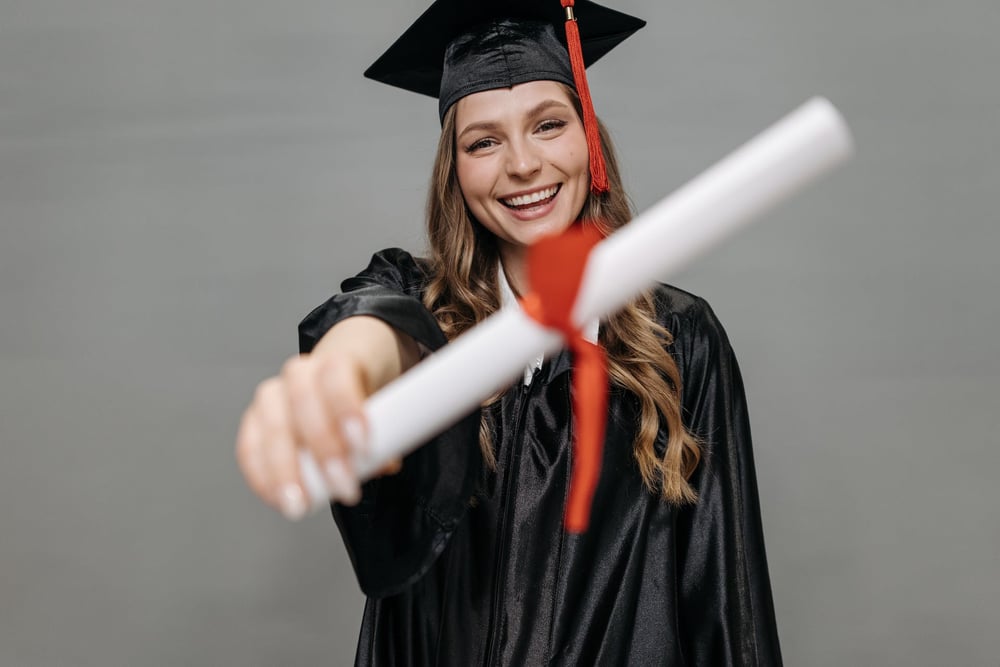 A smiling graduate in a cap and gown holds out a rolled-up diploma.