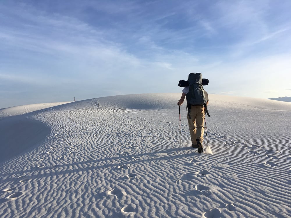 A backpacker carrying trekking poles walks over white sand dunes in a desert.