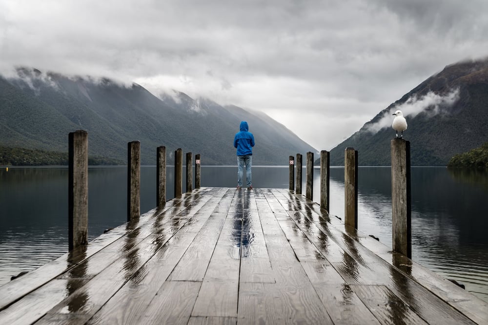 A person standsat the end of a wet dock, looking across a lake at mountains surrounded by fog.