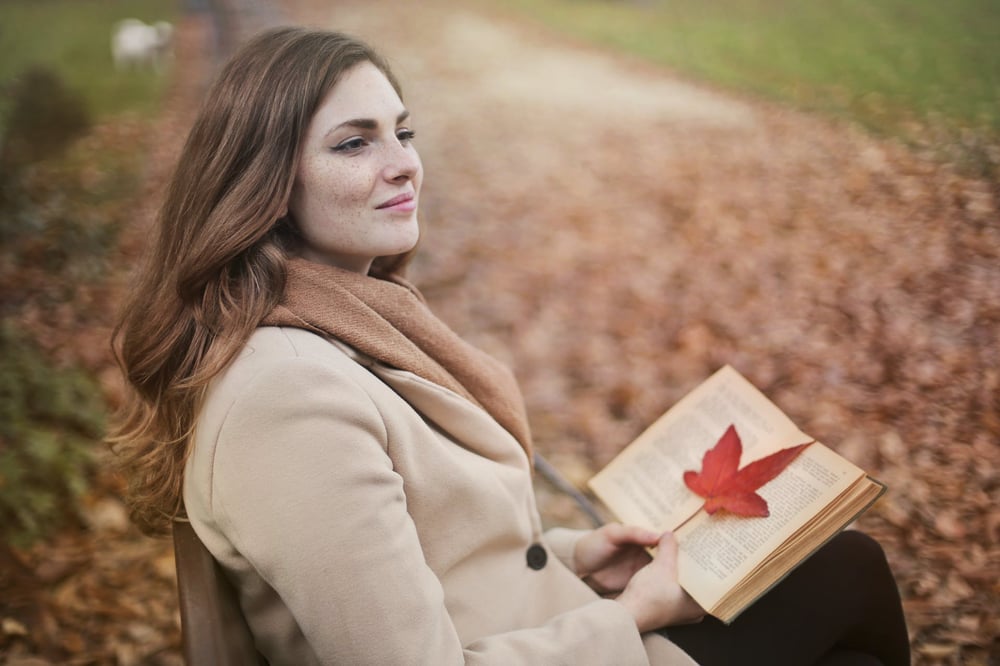 A person in a scarf sits on a chair outside in the fall, holding an open book with a read leaf on the pages.