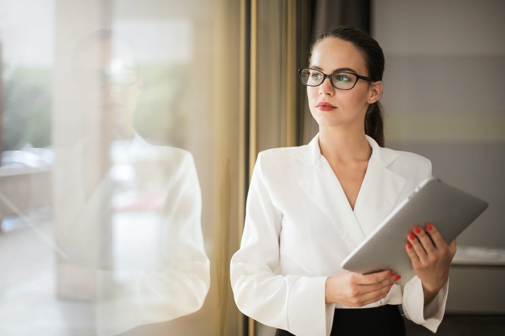 A person in glasses and business attire stands looking out of a window.