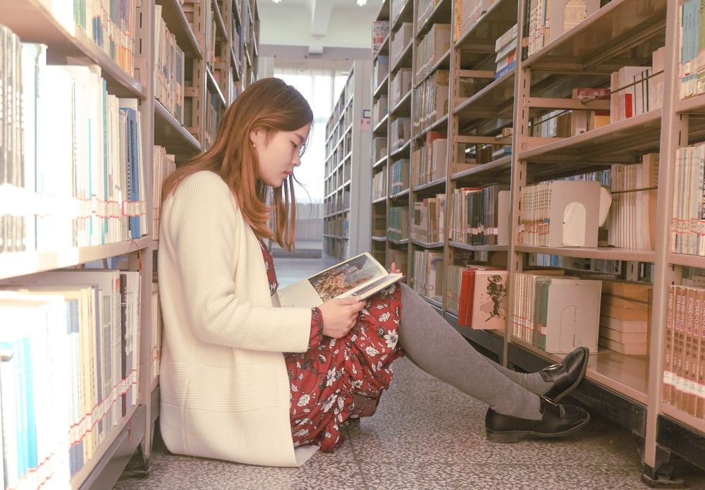 A person sits on the floor, leans against a library bookshelf, and reads a book.