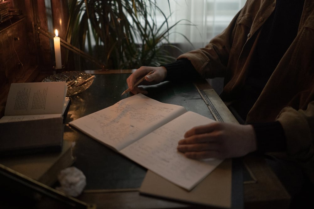 Hands rest on an open notebook beside a candle on a desk in a dim room.