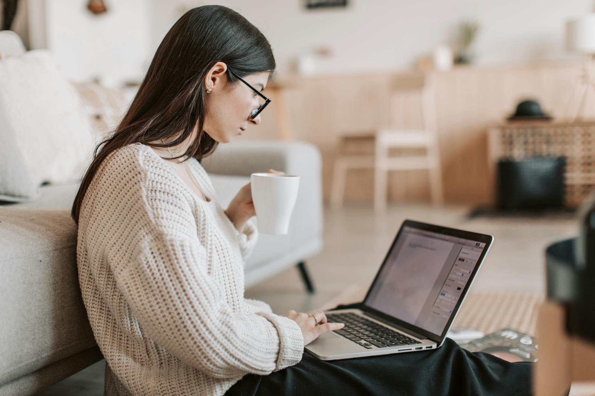 A writer sits on the floor against a couch, holding a cup of coffee and typing on a laptop.