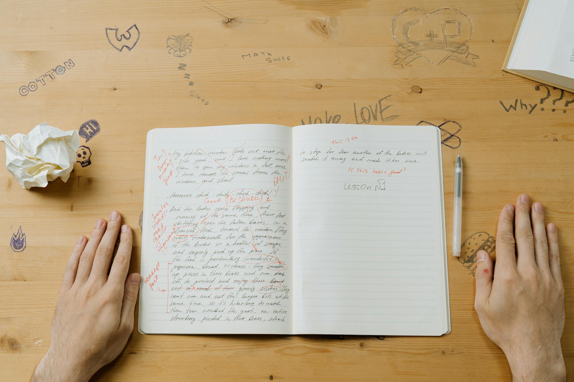 Two hands rest on a desk on either side of an open notebook marked up in red pen.