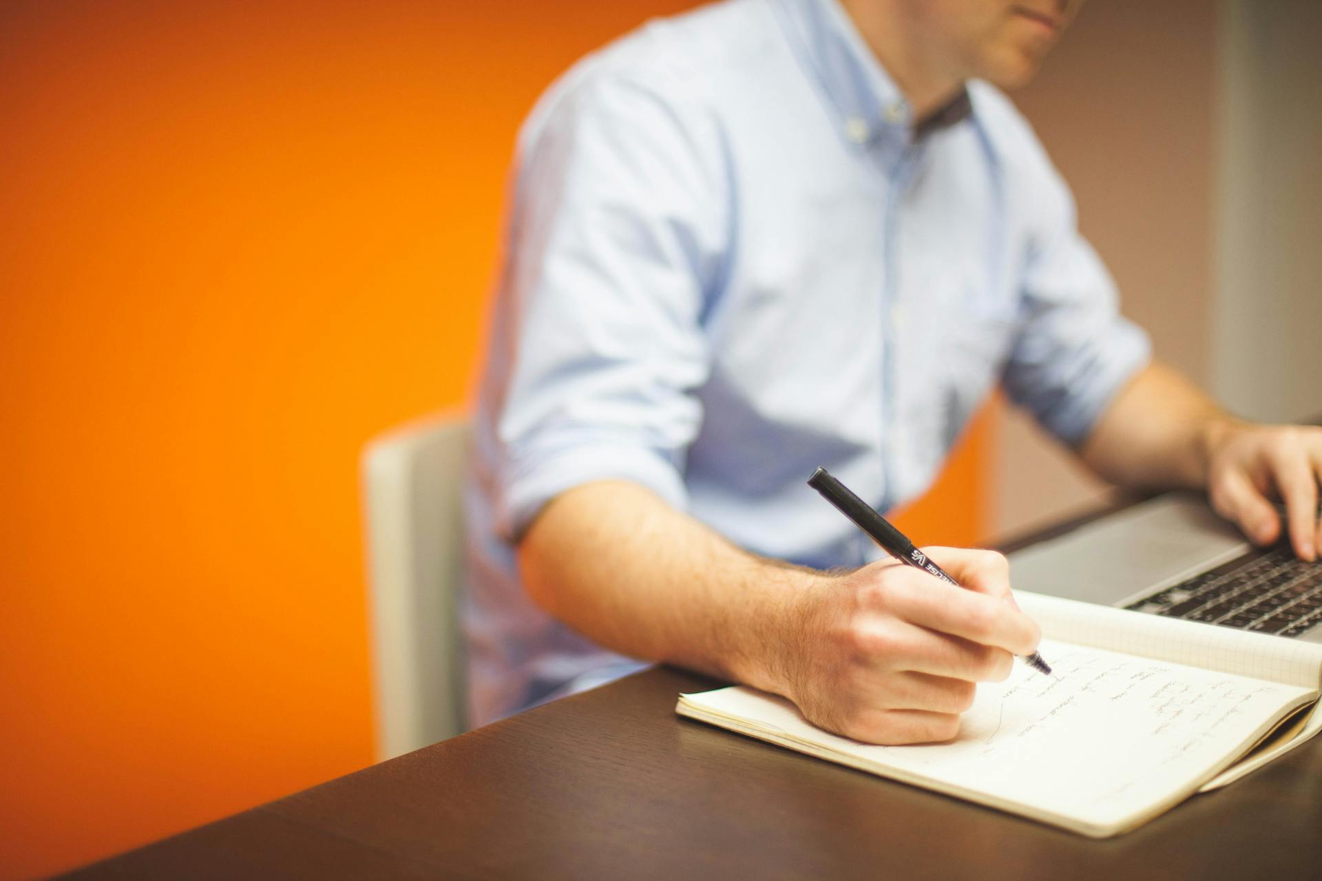 A person sitting in front of an open laptop writes in a notebook.