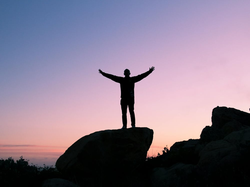 Silhouette of a person standing on a rock with their arms outstretched.