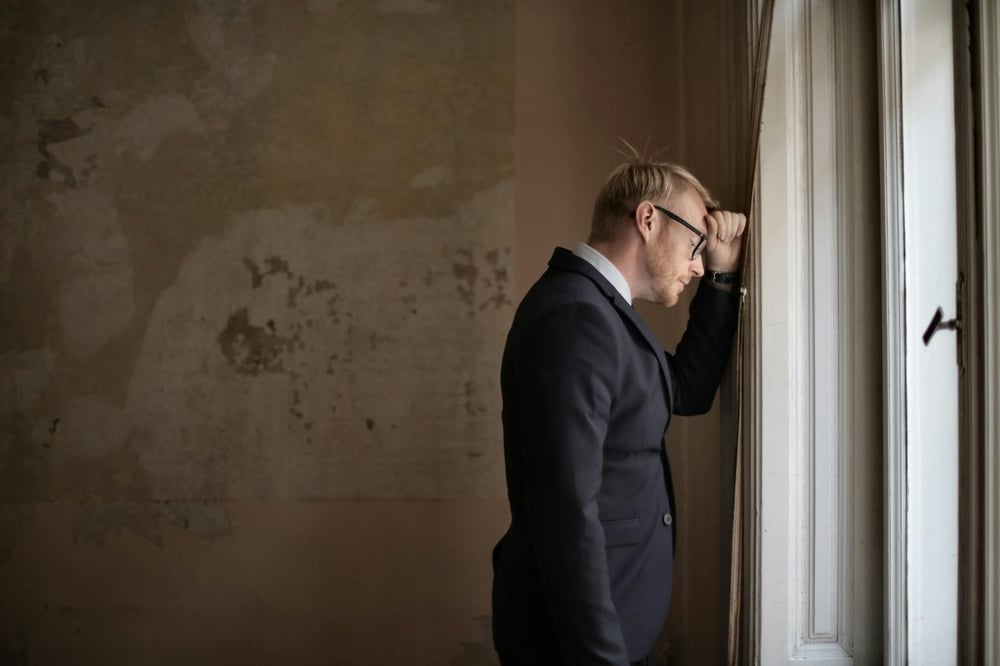 An upset person in a suit rests their head against a wall beside a window.