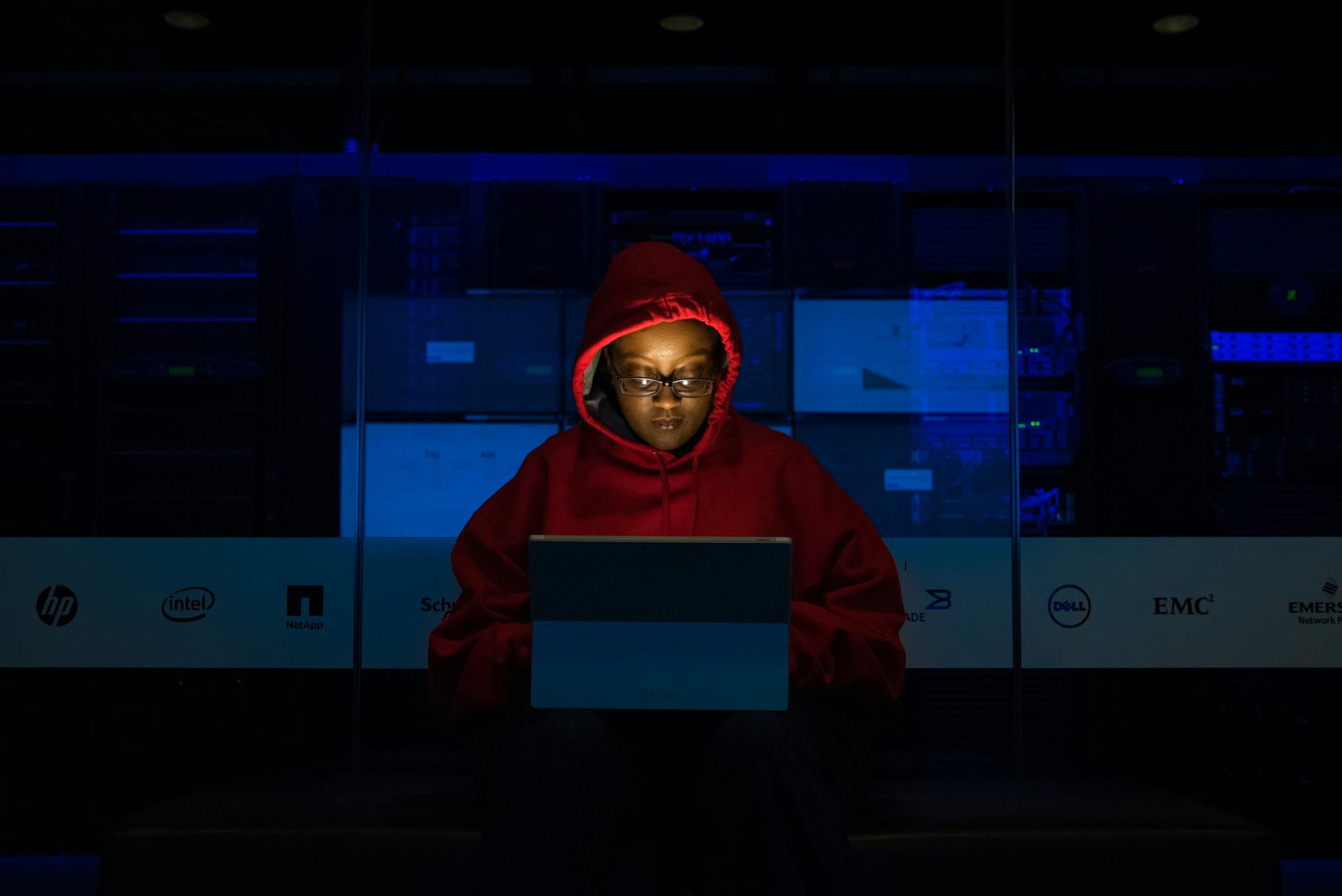 A writer in a red hoodie types on a laptop in a dark room.