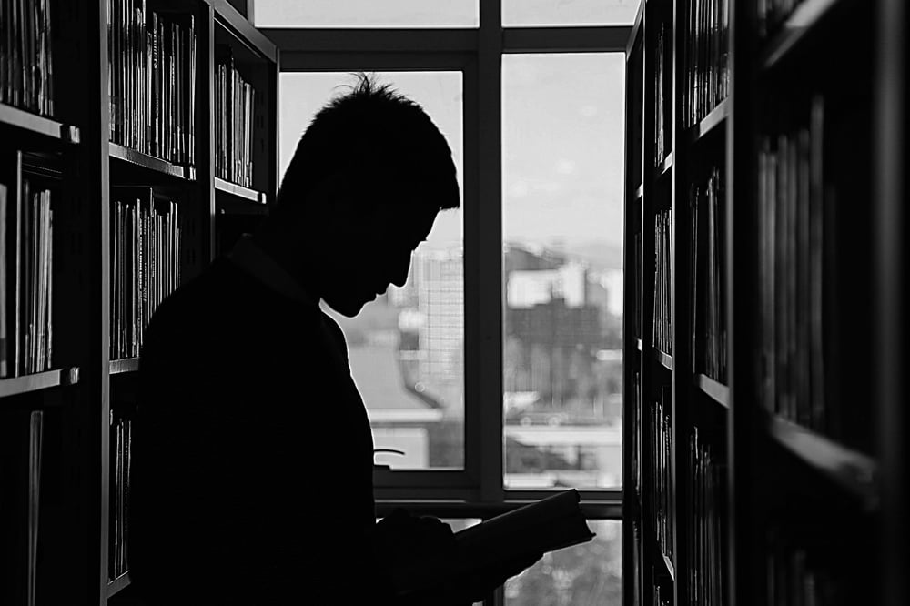 Silhouette of a person standing in front of a window, reading a book between two library bookshelves.
