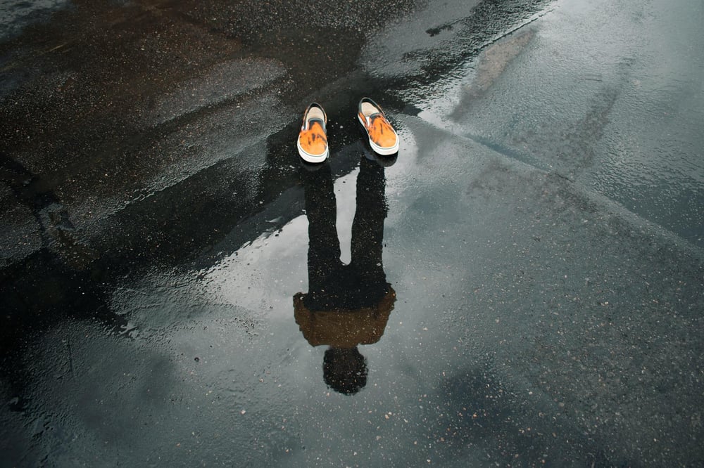A person's reflection appears on wet asphalt but only a pair of empty shoes are actually there.